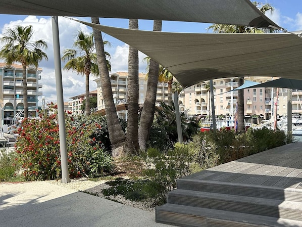 Um calçadão costeiro com palmeiras, um deck de madeira e um porto com barcos ao fundo.