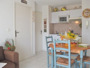 Uma sala de jantar com mesa de madeira e cadeiras azuis, uma porta branca e uma cozinha com micro-ondas e objetos decorativos.