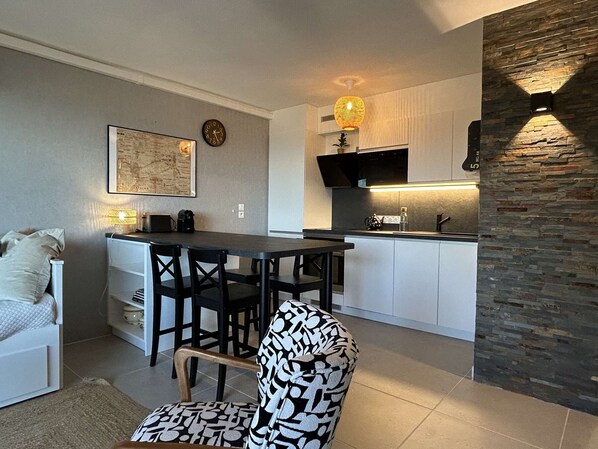 A modern kitchen with white cabinets, a black countertop, and a stone accent wall. There is a dining area with a table and chairs, and a small seating area with a sofa and a patterned armchair.