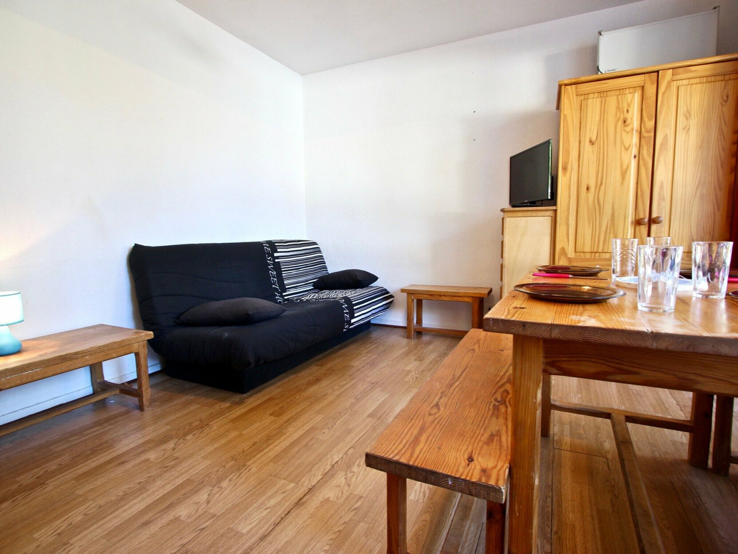 Una habitació petita i acollidora amb un sofà negre, una taula de dinar de fusta i un moble de fusta amb un televisor.