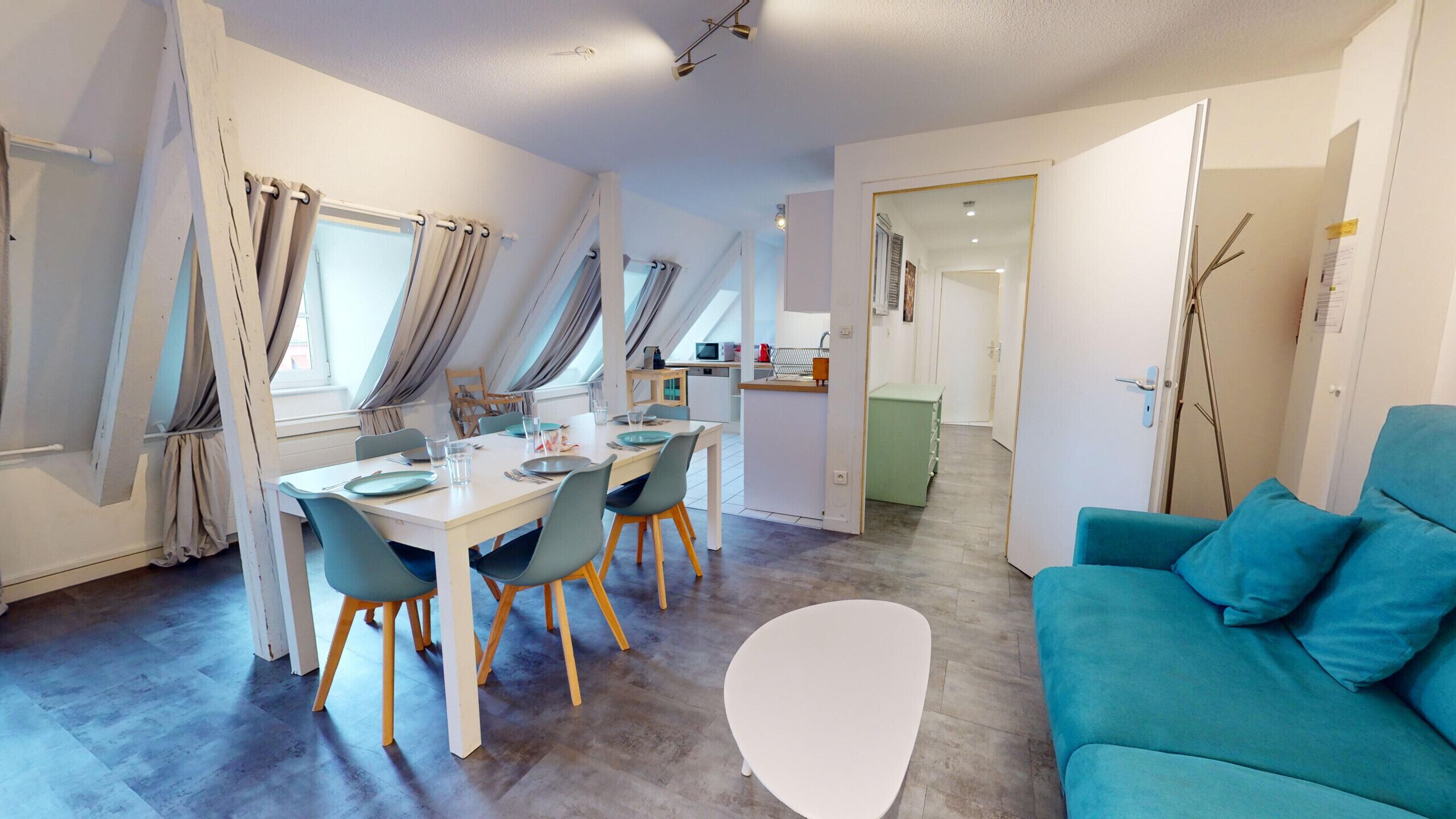 Een moderne woonkamer met een blauwgroene bank, een eethoek met een witte tafel en stoelen, en een keuken op de achtergrond.