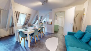 Una sala de estar moderna con un sofá color turquesa, un comedor con una mesa y sillas blancas, y una cocina al fondo.