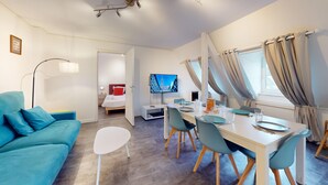 Una habitación de hotel moderna con un sofá azul, una mesa de comedor con sillas, un televisor y una cama en la habitación contigua.