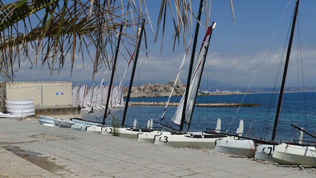 A waterfront with sailboats docked along the shore, a building with a sign, and a clear view of the sea and distant buildings. - Deluxe garden level apartment, close to beaches, between Salis and Pinewoods (Antibes)