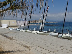 A waterfront with sailboats docked along the shore, a building with a sign, and a clear view of the sea and distant buildings. - Deluxe garden level apartment, close to beaches, between Salis and Pinewoods (Antibes)