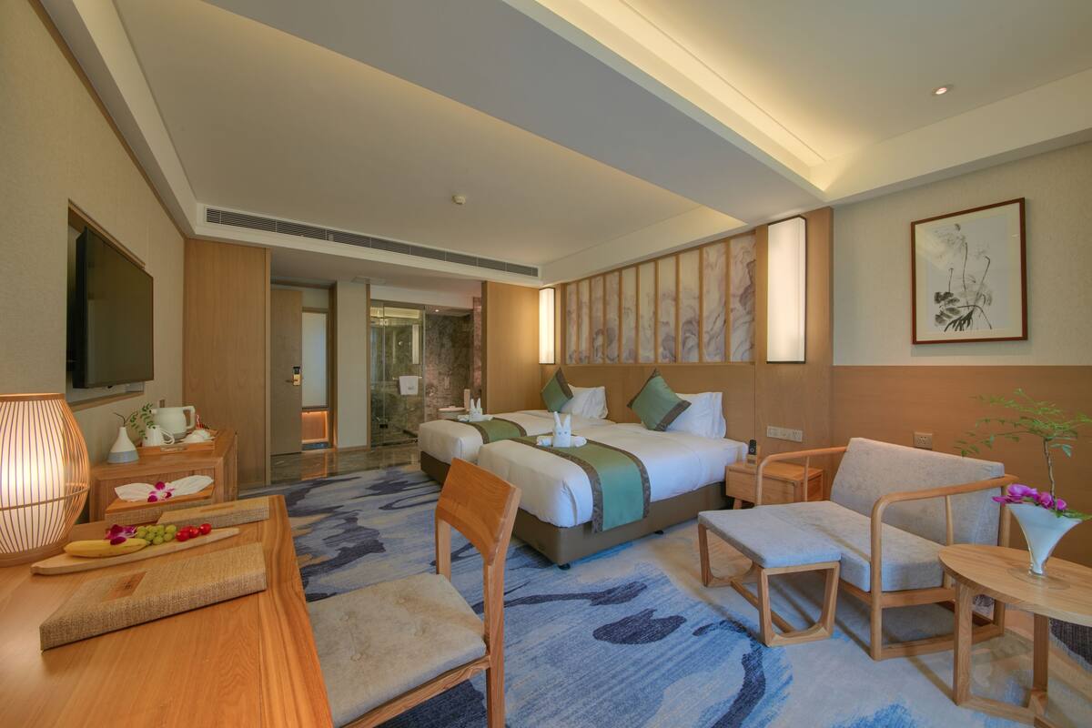Una habitación de hotel moderna con una cama grande, dos mesitas de noche, una zona de estar con sofá y sillones, una pequeña mesa con un florero y obras de arte en las paredes.