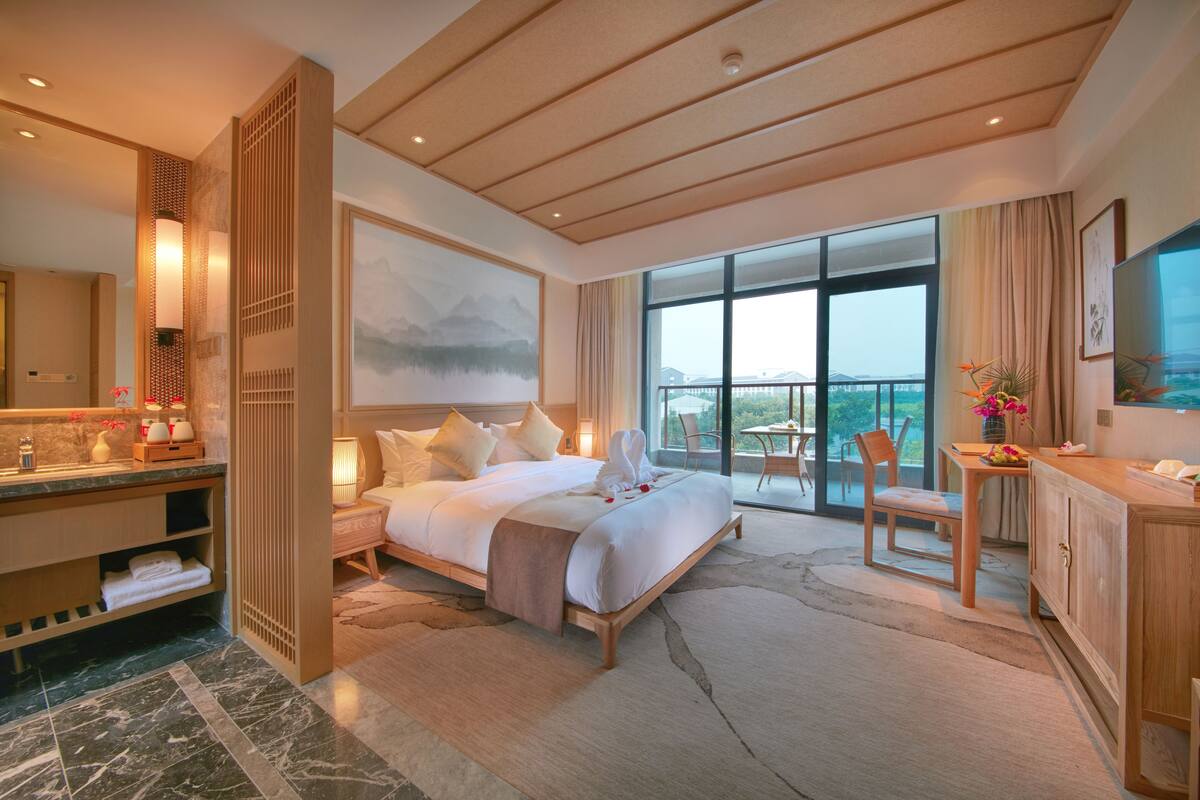 Un dormitorio amplio con una cama grande, una zona de estar con una mesa y sillas, un balcón con vistas y un baño con tocador y ducha.