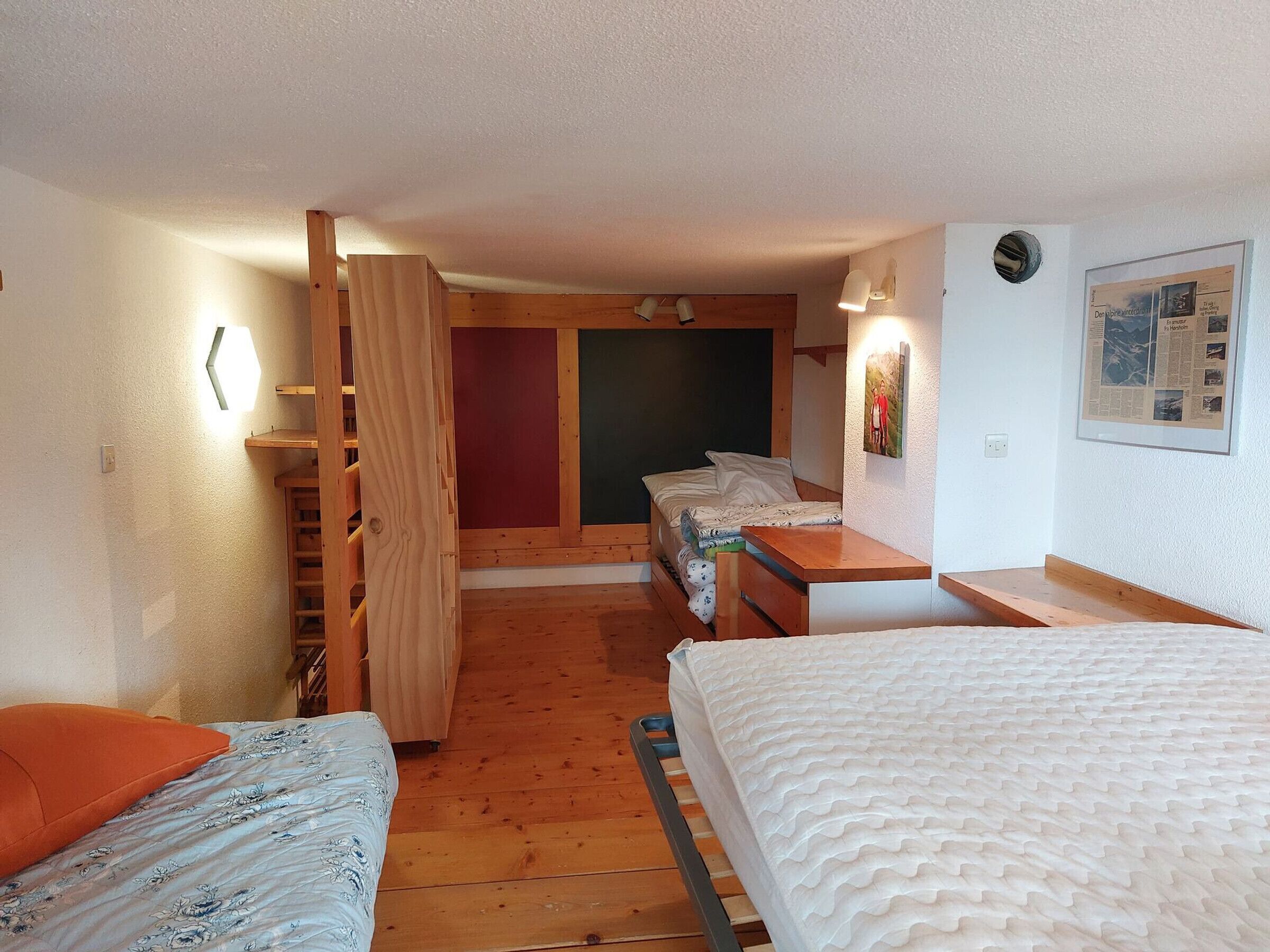 A compact room with a bed, a desk, and a chair. There is a wooden ladder and a small table.