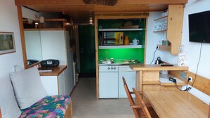 Une cuisine compacte avec un revêtement mural vert, une table à manger en bois et un canapé agrémenté d’un coussin coloré.