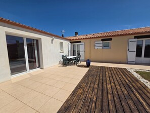 A patio with a table and chairs, a wooden deck, and a building with a tiled roof and sliding glass doors.