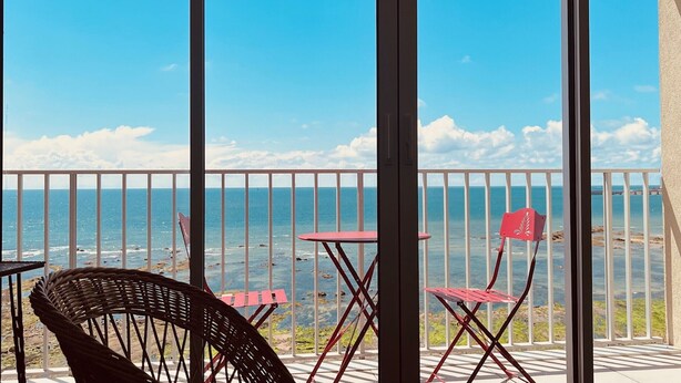 A balcony with a view of the sea, featuring a wicker chair, a red folding chair, and a small table.