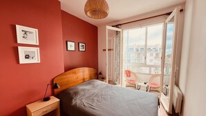 A bedroom with a wooden headboard, a single bed, a nightstand, a lamp, and a large window with a view of buildings.