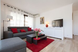 A modern living room with a grey sofa, red cushions, a coffee table, and a red rug. A flat-screen TV is mounted on the wall, and there is a vase with pink flowers on the coffee table.