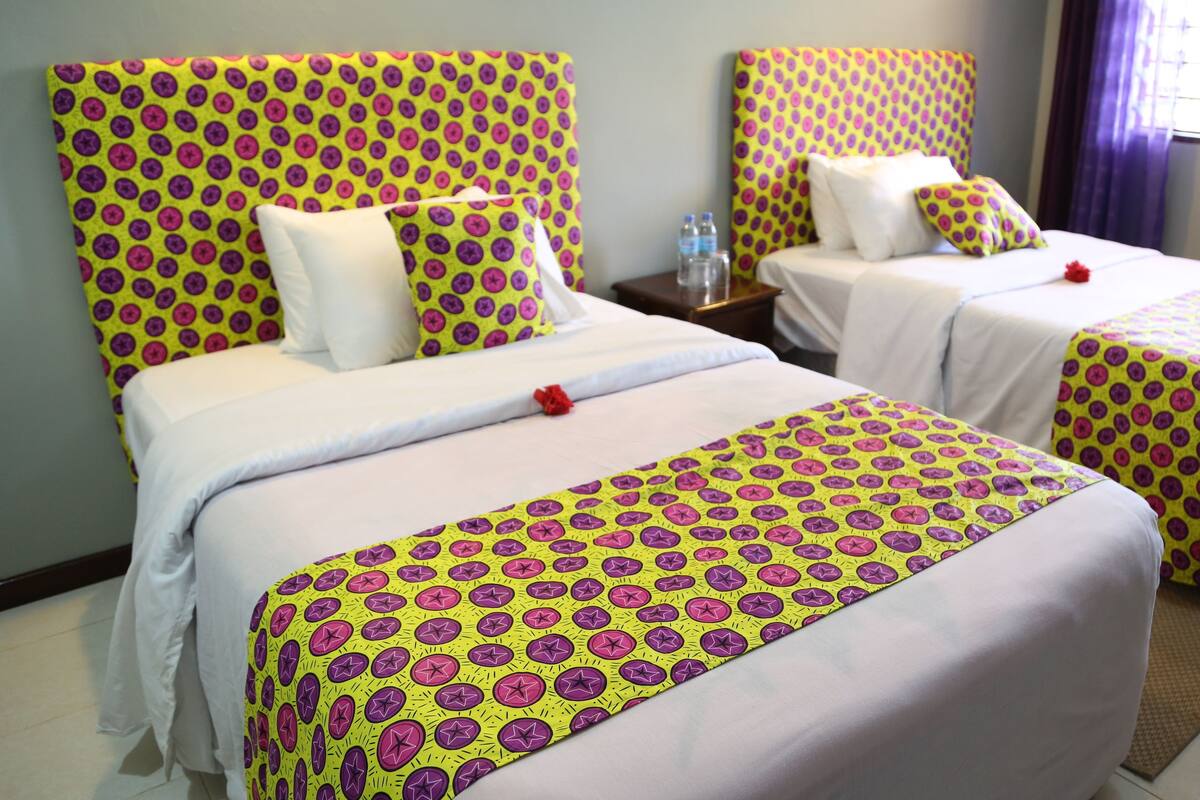 Quarto de hotel com duas camas de solteiro, cada uma com colcha estampada e almofadas combinando. Há uma mesa de cabeceira com uma garrafa de água e uma luminária entre as camas.