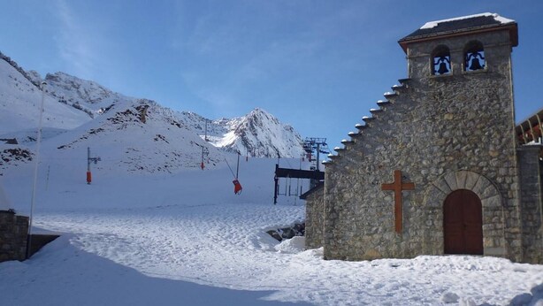 Una capilla de piedra con una cruz, rodeada de montañas nevadas y equipo de telesilla.