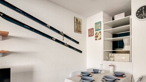 Un comedor con una mesa dispuesta para cuatro personas, sillas blancas, un reloj en la pared y estanterías con diversos objetos.