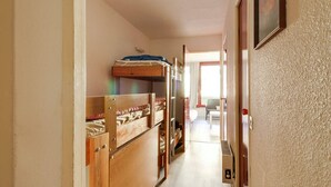 Un pasillo estrecho con literas a un lado y una puerta que da a una habitación con cama y sillón.