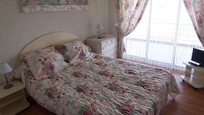 Een slaapkamer met een bed, nachtkastje, dressoir, televisie en raam met gordijnen.