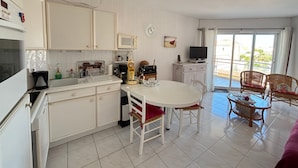 Een compacte keuken met witte kasten, een eettafel met stoelen, een televisie en een balkon met rieten stoelen.