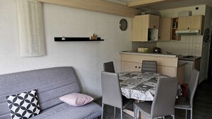 A compact living space with a grey sofa, a dining table with four chairs, a kitchen area with wooden cabinets, and a wall-mounted clock. - Ski-in/Ski-out Apartment for 6 with Balcony, South-Facing, 1 Bedroom, La Mongie (La Mongie)