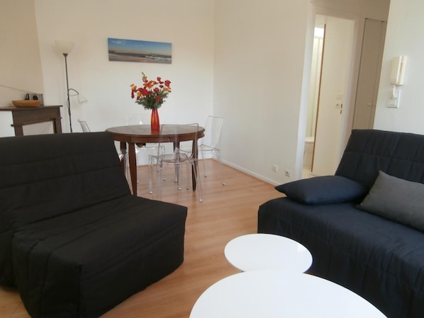 A living room with two black sofas, a white coffee table, and a dining table with clear chairs. A vase with flowers is on the dining table.