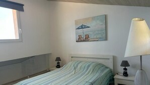 Une chambre à coucher avec un lit, un tableau représentant des chaises de plage et un parasol, une lampe et une fenêtre avec un rideau bleu.