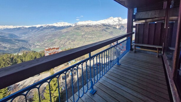 Een houten balkon met een blauwe reling met uitzicht op een bergachtig landschap met besneeuwde toppen.