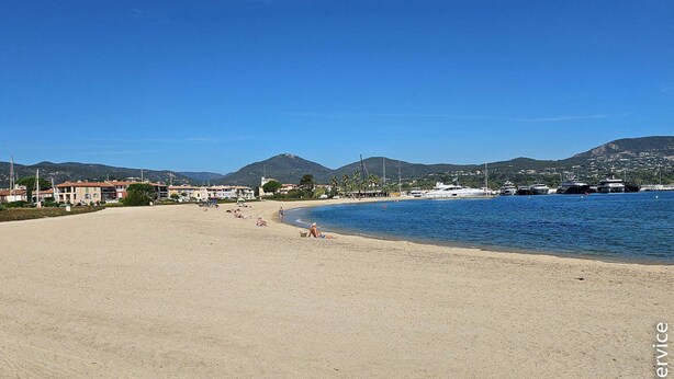 Une plage avec une eau d’un bleu limpide, un rivage sablonneux et des bâtiments en arrière-plan.
