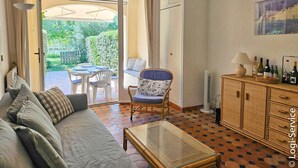 Un salon confortable avec un canapé, une table à manger et une chaise. On y trouve une table basse en verre et un meuble contenant des bouteilles. La pièce offre une vue sur un jardin grâce à une porte ouverte.
