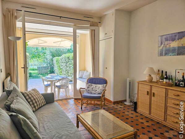 Un salon confortable avec un canapé, une table à manger et une chaise. On y trouve une table basse en verre et un meuble contenant des bouteilles. La pièce offre une vue sur un jardin grâce à une porte ouverte.