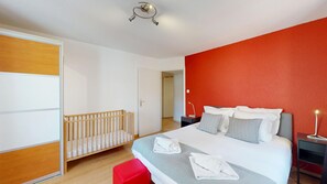 A bedroom with a crib, a bed with pillows, a nightstand, and a red accent wall. - schlumberger 120m2 city center 3br 2bth (Colmar)