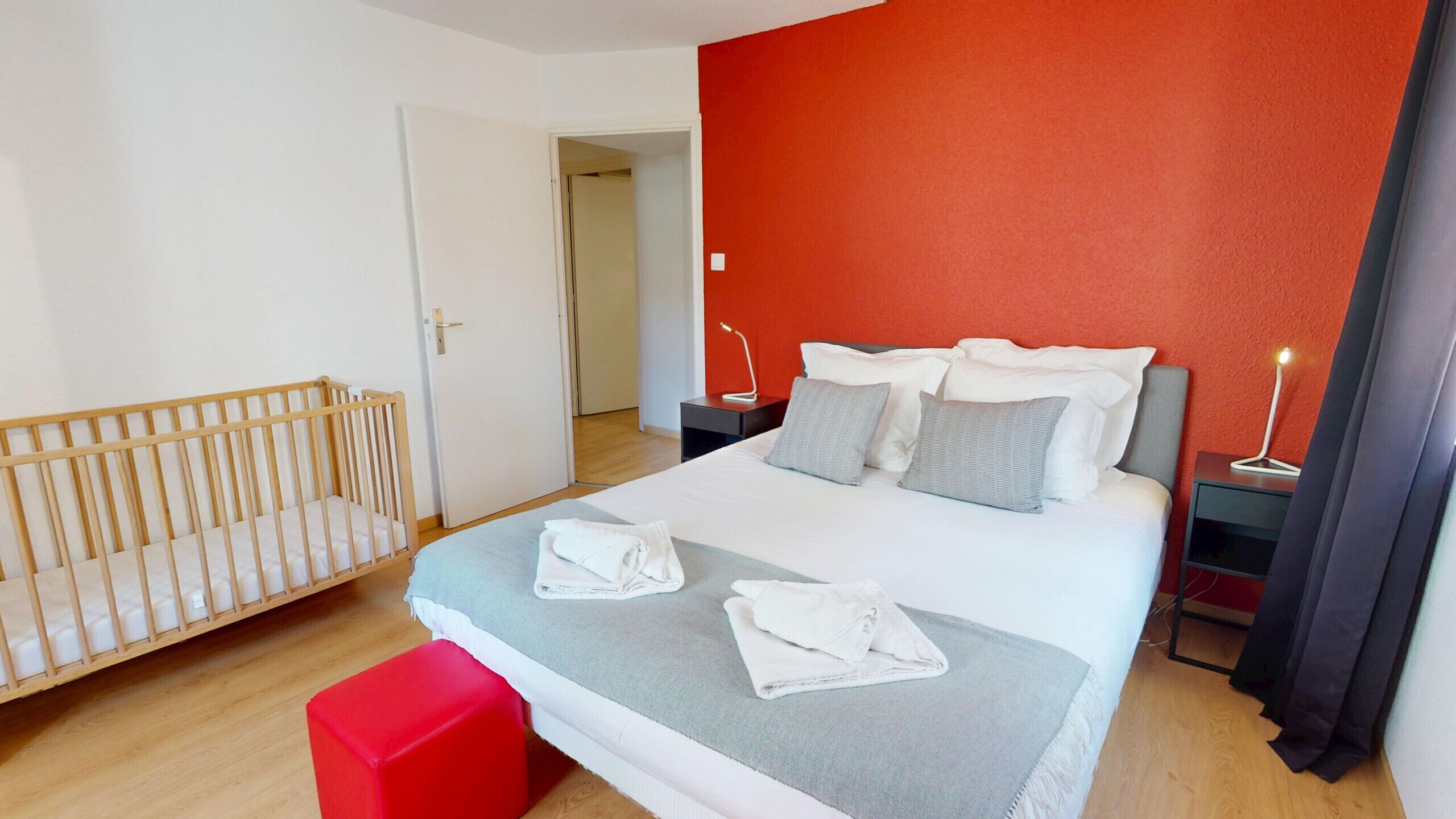 Une chambre à coucher avec un lit, un berceau, un pouf rouge et une table de chevet avec une lampe.