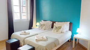 A double bed with two pillows, a bedside table, a window with curtains, and a view of buildings outside.
