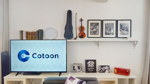 A room with a television displaying 'Cotoon,' a bookshelf with books, a guitar, framed pictures, and a small table with a box. - schlumberger 120m2 city center 3br 2bth (Colmar)