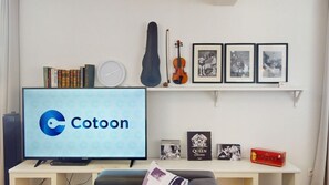 A room with a television displaying 'Cotoon,' a bookshelf with books, a guitar, framed pictures, and a small table with a box.