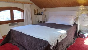 Una camera da letto con soffitto in legno, un letto con copripiumino bianco e marrone scuro, un comodino con lampada e una finestra con tende.
