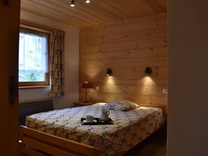 Un dormitorio con techo de madera, una cama con ropa de cama floral, una ventana con cortinas estampadas y dos luces fijadas a la pared.