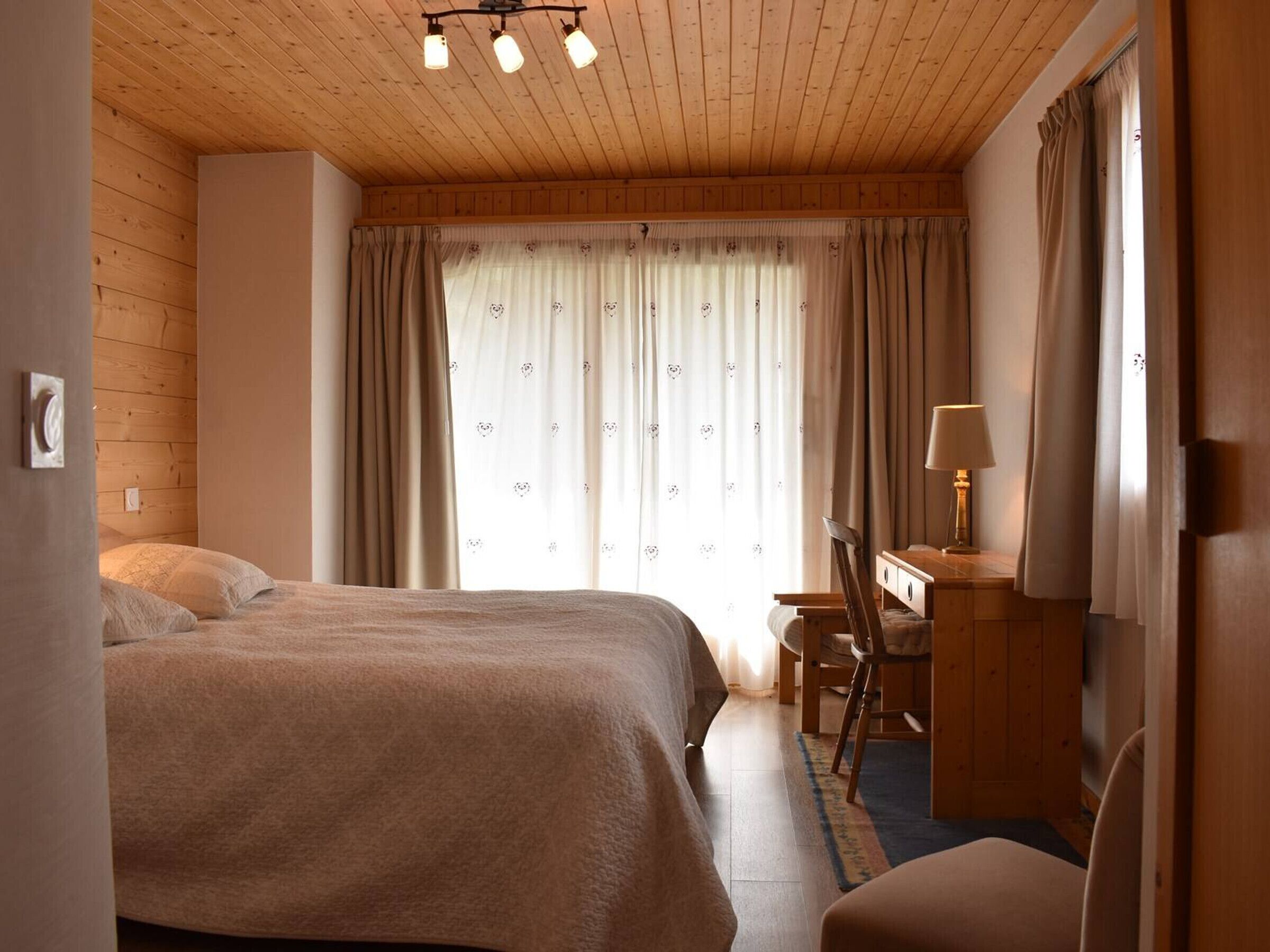 A cozy room with a wooden ceiling, a bed with a beige blanket, a desk with a lamp, and a chair.