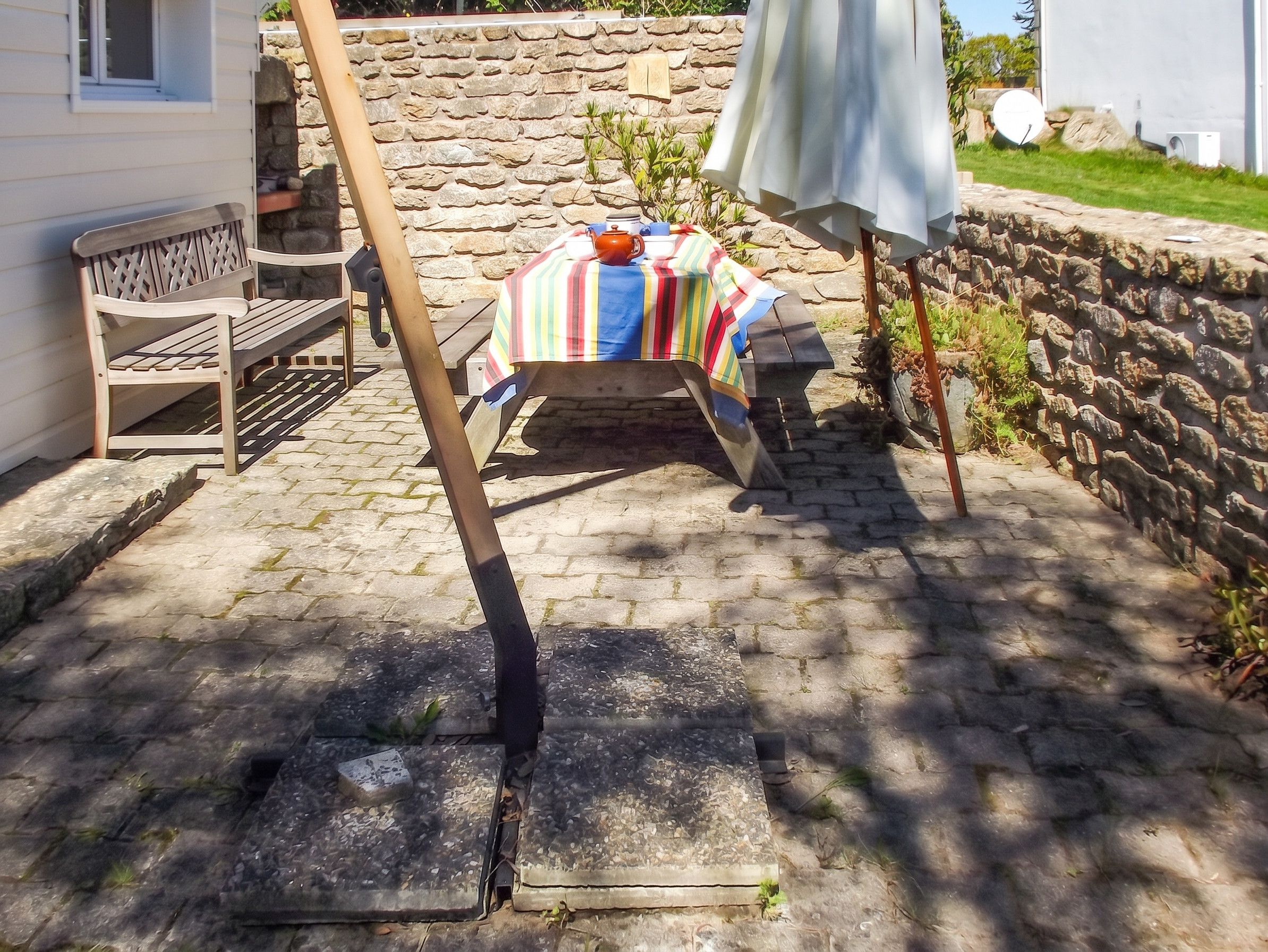 Un mur de pierre, un banc en bois, une table recouverte d’une nappe à rayures et un parasol blanc.