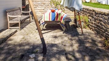 Un mur de pierre, un banc en bois, une table recouverte d’une nappe à rayures et un parasol blanc.