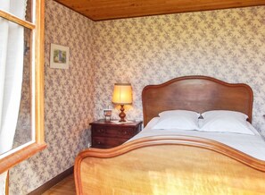 A bedroom with a wooden bed, a nightstand with a lamp, floral wallpaper, and a window with curtains. - Homerez - Charming House in Trédrez-Locquémeau + Garden (Trédrez-Locquémeau)