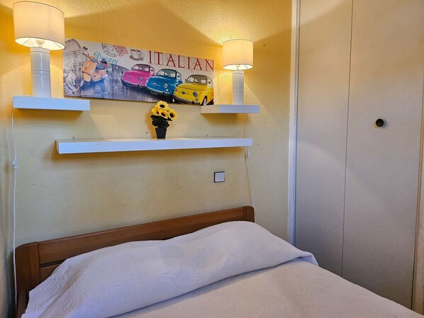 A bedroom with a bed, a wall-mounted shelf with a lamp, and a wall decoration featuring vintage cars.