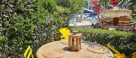 A round wooden table with woven placemats, yellow chairs, and a lantern, set outdoors with greenery and boats in the background.