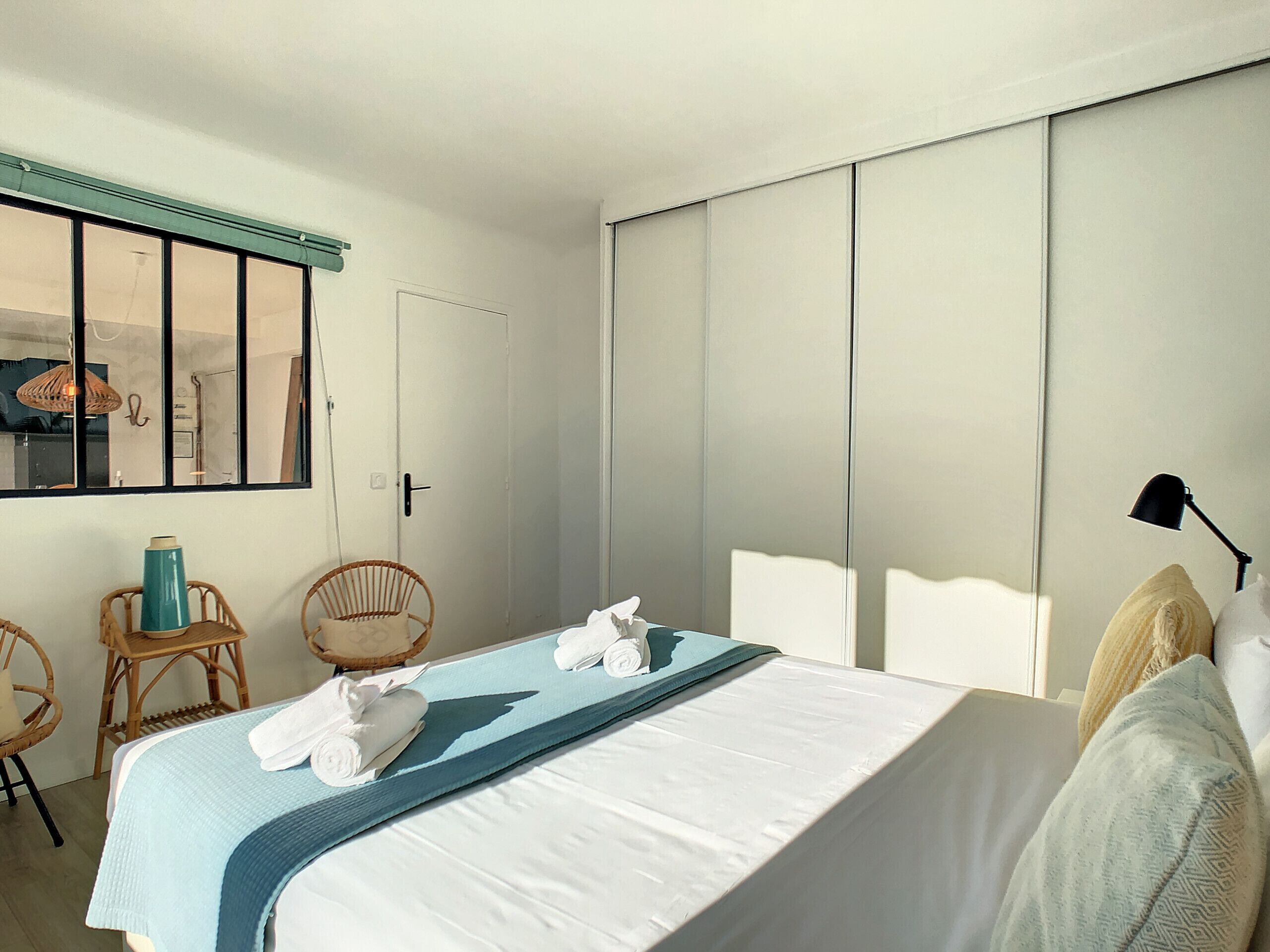 Habitació d'hotel amb un llit, dues cadires de vimetla, una tovallola blava i una làmpada.