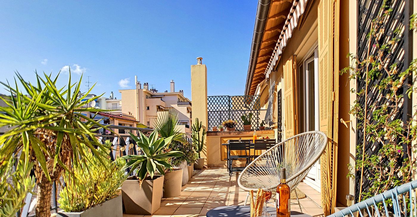 A rooftop terrace with potted plants, a wicker chair, and a table with a bottle and glasses.
