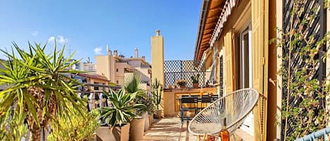A rooftop terrace with potted plants, a wicker chair, and a table with a bottle and glasses.