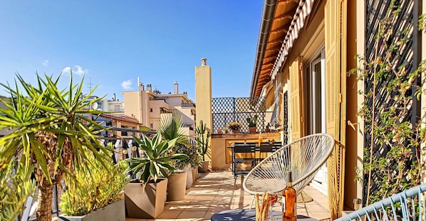 Eine Dachterrasse mit Blumentöpfen, einem Korbstuhl und einem Tisch mit Flasche und Gläsern.