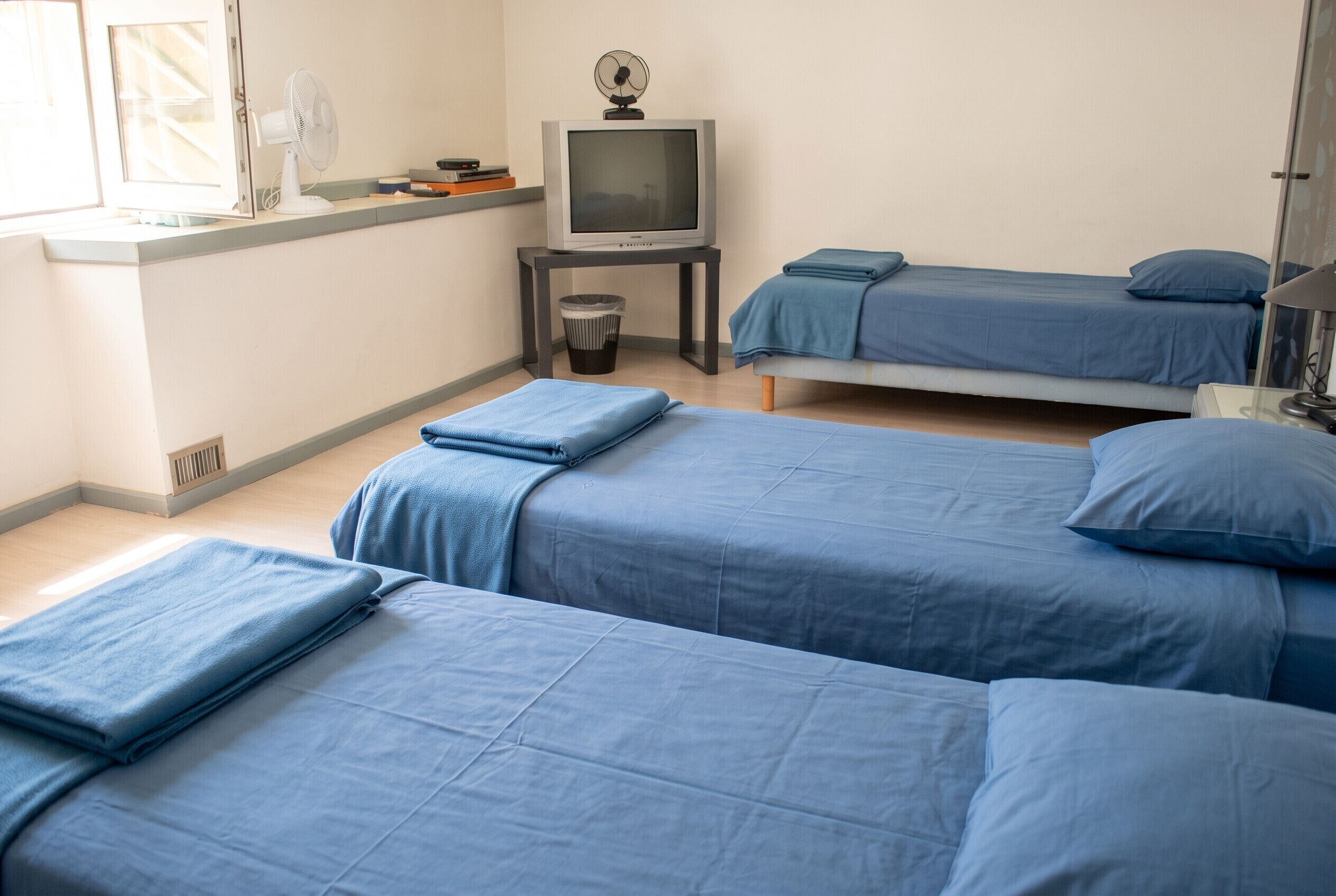 Ein Zimmer mit drei Betten, einem Fernseher, einem Ventilator und einer Mülltonne.