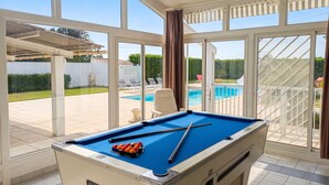 Une table de billard avec des billes, une chaise et une vue sur une piscine à travers de grandes fenêtres.