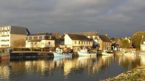 A riverside town with buildings along the water, boats docked, and a calm river reflecting the structures. - Ground floor apartment with terraces, WIFI, parking, and pets allowed (Barneville-Carteret)